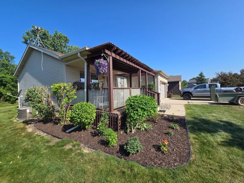 Small house with covered porch and hanging flowers, surrounded by landscaped yard and parked vehicles on a sunny day
