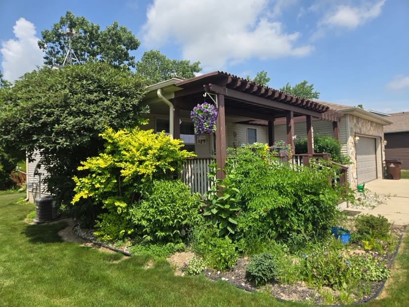 Brown house with covered porch surrounded by lush green landscaping, flowering shrubs, and trees under a blue sky